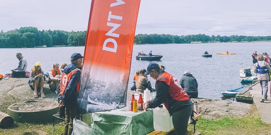 En gjeng med mennesker er samlet ved vannet, i front er det et DNT Vansjø beachflagg.