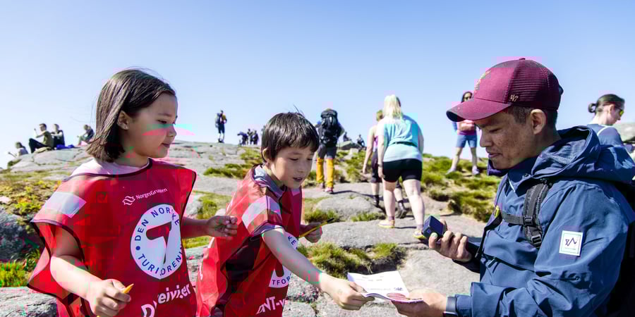 En jente og en gutt med rød T-vest deler ut skjema til en turgåer under et stort arrangement på en fjelltopp i Rogaland.