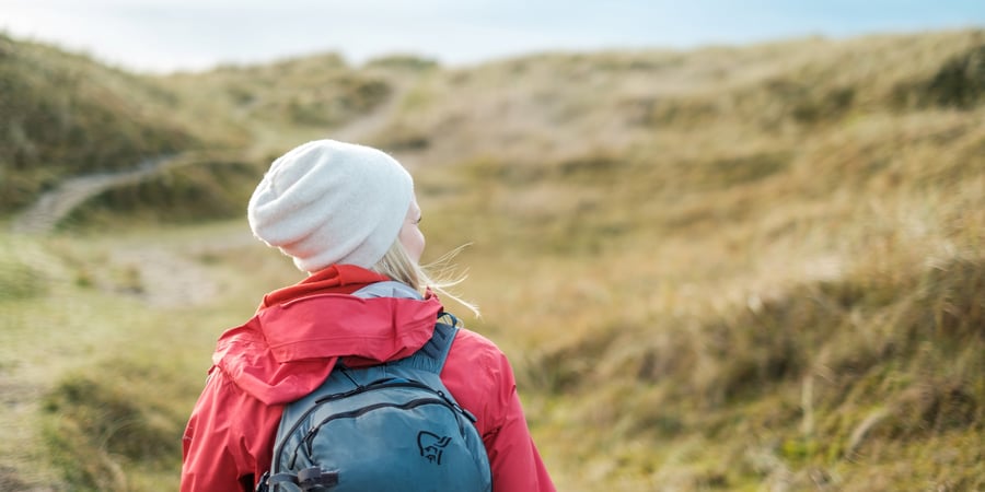 En kvinne med sekk, rød jakke og lue går nedover sand-dynene på Jæren.
