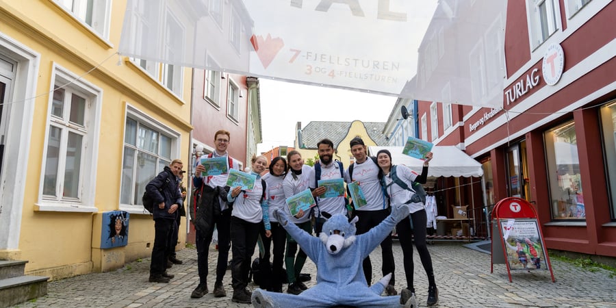 Maskot sitter foran gruppe mennesker i bygater og jubler.