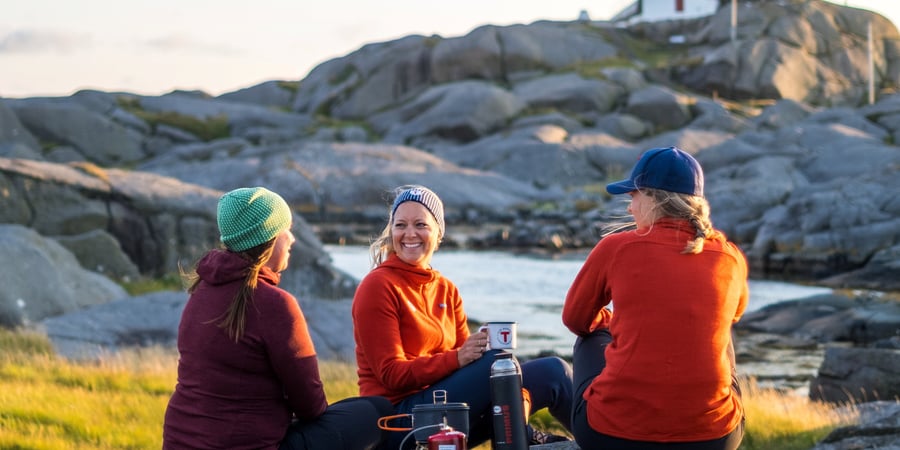 Tre damer sitter på et berg og snakker sammen. Et fyrtårn vises i bakgrunnen.