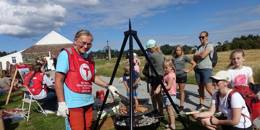 En kvinne med rød T-vest står ved en bålpanne og smiler til kamera. Mange folk rundt henne som er på et arrangement og skal grille pølser på bålpanna.