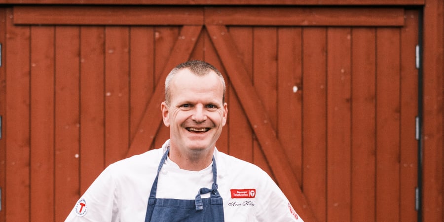 Mann stående med kokkeforkle foran rød låve