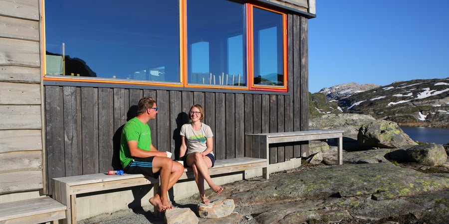 To personer sitter ved hyttevegg i sol og prater.