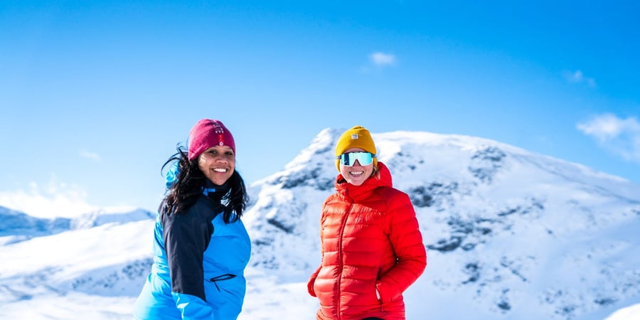 To damer ser inn i kameraet og smiler. Ei har på seg blå jakke, mens hun andre har oransj jakke. I bakgrunnen er det snødekte fjell.