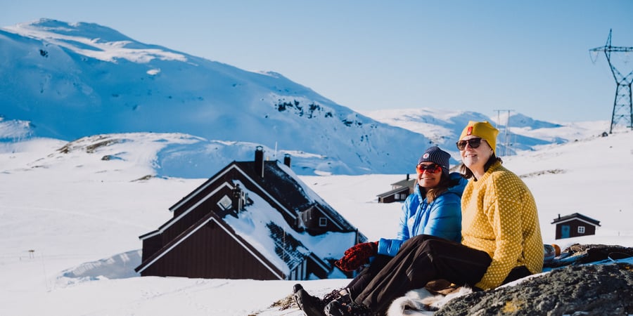 to personer sitter på en steinur og det er masse snø rundt dem. Det er sol og blå himmel, og personene smiler. Man ser ei hytte i bakgrunnen. 