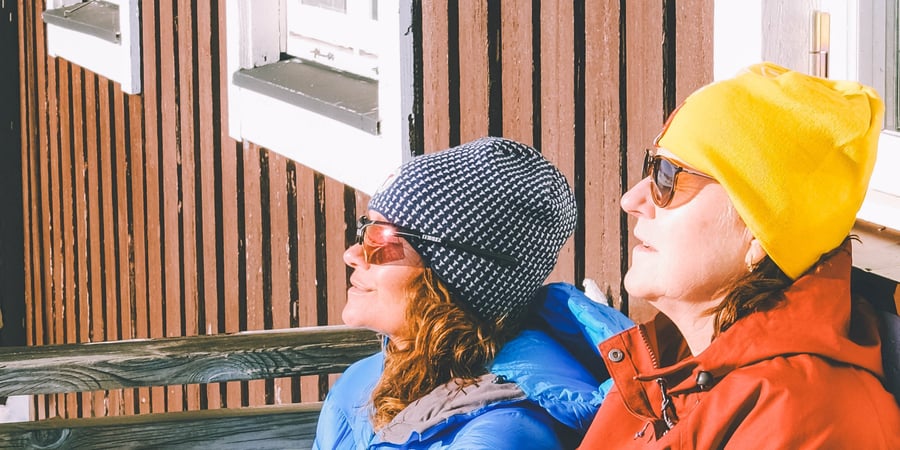 To personer som sitter i solveggen på et hus. Noe snø i bakgrunnen.