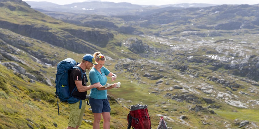 En mann og en dame i shorts og med ryggsekk studerer kart mens de er på tur i fjellet en sommerdag.