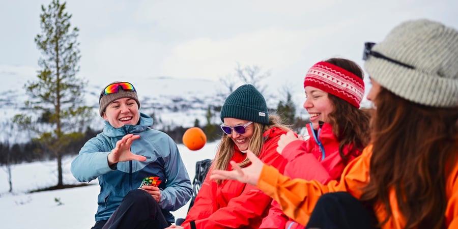 Fire kvinner sitter i vinterlandskap og smiler. Hun ene kaster en appelsin mot hun andre.