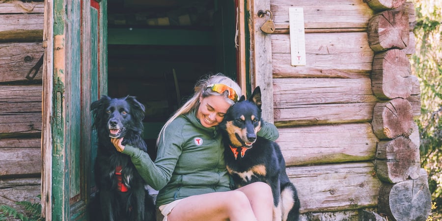kvinne som sitter i døråpningen til en liten tømmerhytte, med en hund på hver side