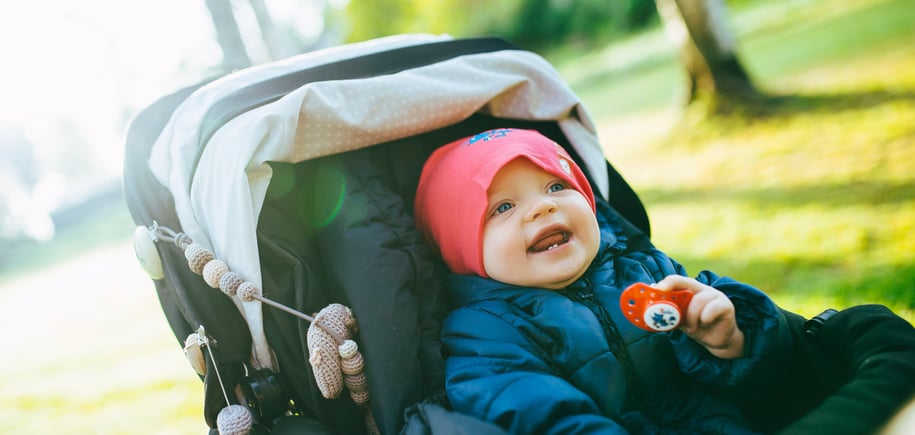En baby med rosa lue og blå jakke sitter i en barnevogn utendørs, smiler og holder i en smokk, med sollys i bakgrunnen.