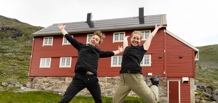 En mann og en dame som hopper med armene i været foran en rødmalt hytte
