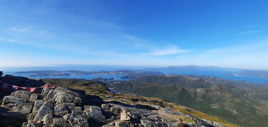Bildet er tatt fra det som ser ut som en fjelltopp, med utsikt over skog og hav. Det er blå himmel.