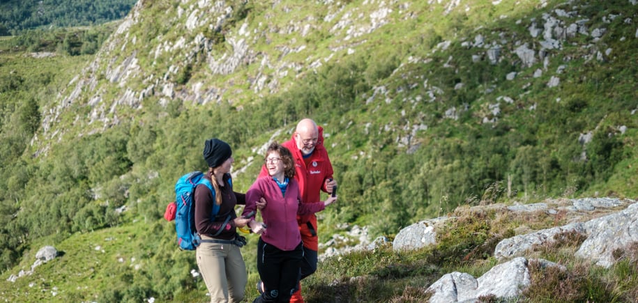 Tre personer går i fjellterreng. Damen i midten har en gul bøyle som hun holder i og blir støttet av to ledsagere. Alle personene smiler og ler.