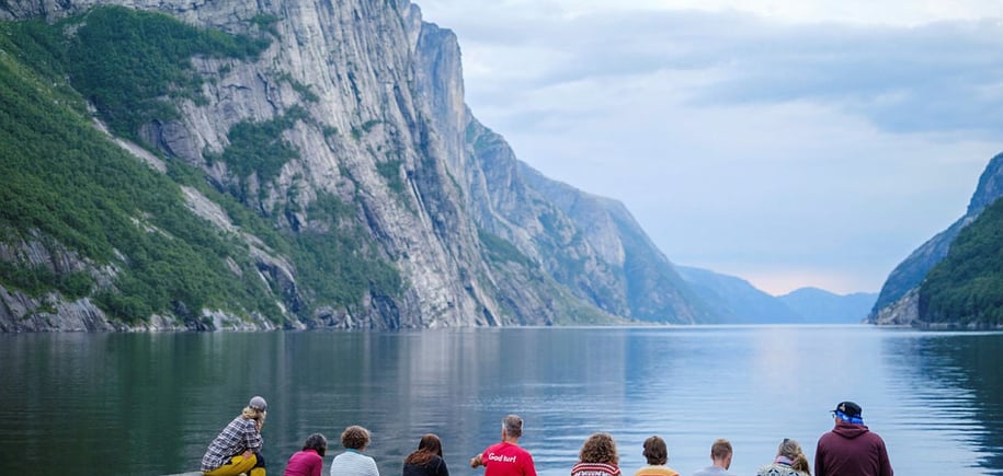 En gruppe mennesker sitter med ryggen til på brygga innerst i en lang fjord.