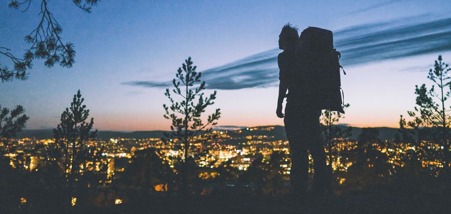 Mann med stor sekk som står og ser utover en opplyst by på kveldstid.