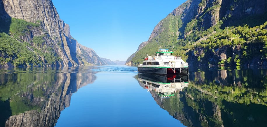 Bilde av rutebåt som kommer kjørende innover fjorden. Speilblank fjord og høye fjellvegger.