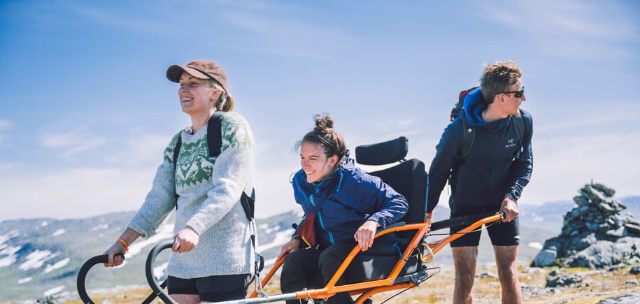 Tre personer ute på tur, der to stykker hjelper den i midten med å holde/bære en modifisert rullestol for bruk ute i naturen. 