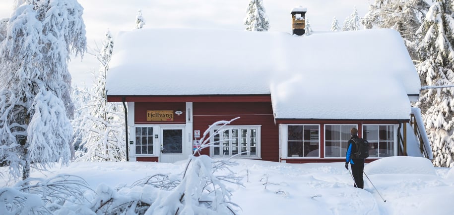 Mann foran en rødmalt hytte med mye snø på taket 