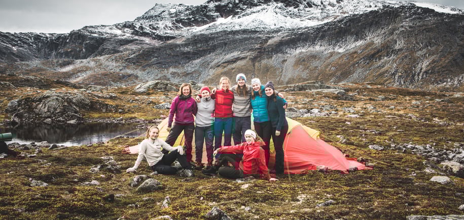 Åtte personer oppstilt foran et telt, to av dem sittende. Alle har på varme klær. Fjell i bakgrunnen.
