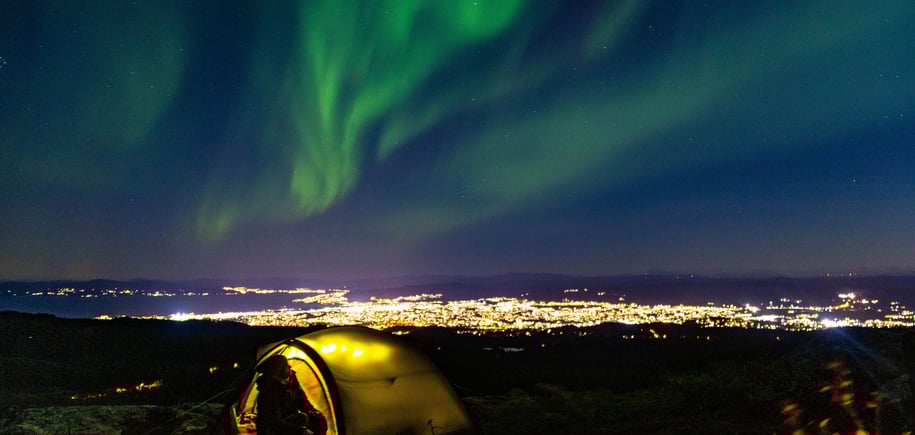 Bildet viser en mann ved et telt på toppen av et fjell, men utsikt over en stor by. Det er natt, og nordlys.