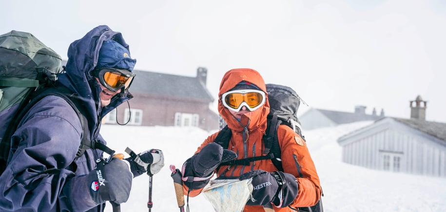 To skiløpere i vinterfjellet, i sterk vind, med hette, briller og kart.