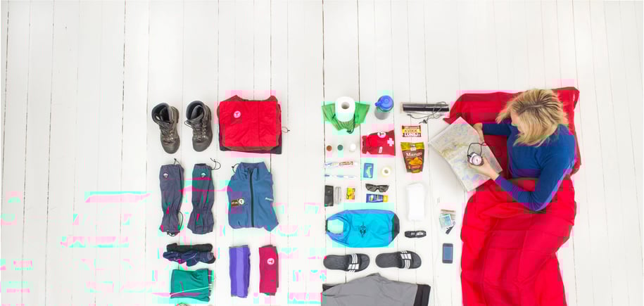 Kvinne ligger i lakenpose med klær og utstyr for fjelltur rundt seg.