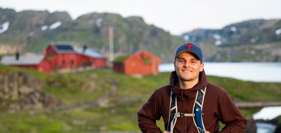 En ung mann med sekk på ryggen ser i kamera. I bakgrunnen vises en rød hytte.