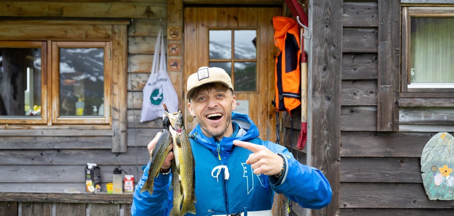 Mann i blå jakke smiler foran hytte og peker på noen fjellørret han har fanget.