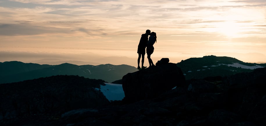 Kjærestepar kysser i solnedgang på fjelltopp.