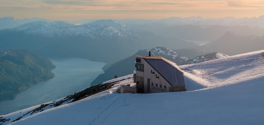 Langstrakt hytte på toppen av fjell i solnedgang med fjord lenger bak.