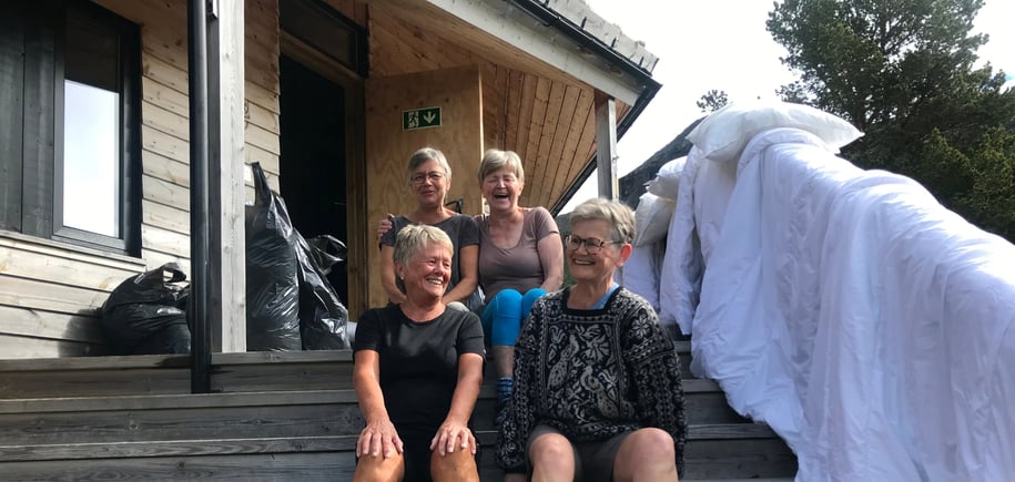 Fire damer sitter på en trapp utenfor en grå hytte. Det henger dyner til lufting.