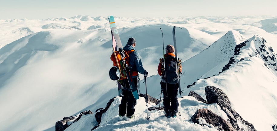 To personer med sekker hvor det er festet på ski på. De står og ser utover et snødekket landskap