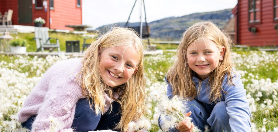 To jenter som sitter på huk og smiler til kamera mens de plukket myrull. En rød hytte i bakgrunnen.