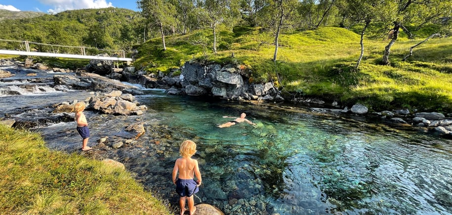 Bading i badekulp, en voksen mann bader og to unge gutter står ved elvekanten og ser på.