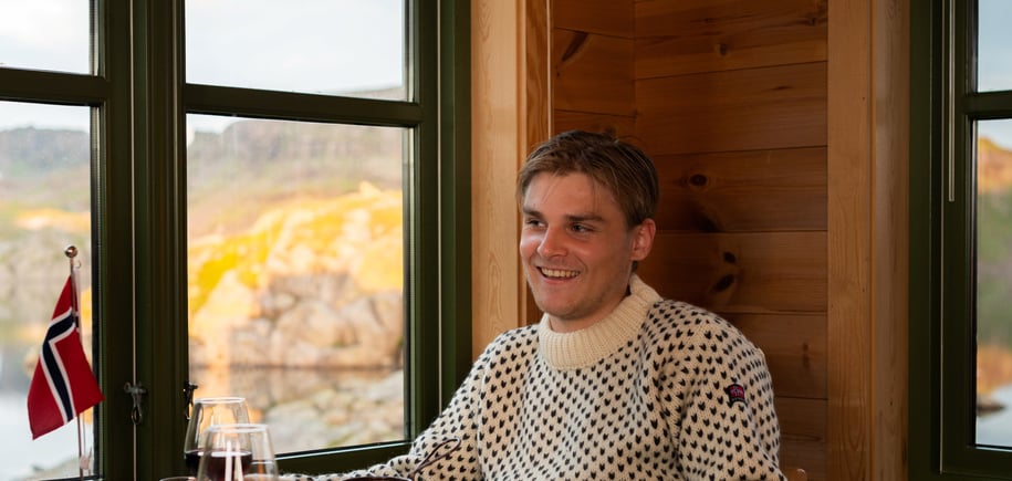 En mann sitter ved et bord og smiler. Foran han er det en tallerken med mat og et norsk flagg.