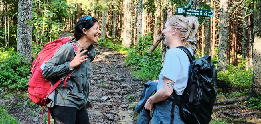 To personer smilende og pratsomme på en skogsti i Oslomarka. Begge bærer tursekker, og i bakgrunnen sees et skilt som viser retningene til Vettakollen (1,6 km) og Sognsvann (3,6 km)