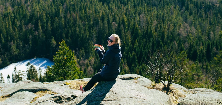 En kvinne sitter på en stein ute i naturen med en skogkledd åsside i bakgrunnen. Hun smiler mens hun holder en Kvikk Lunsj-sjokolade i hånden og har på seg solbriller og en mørk turjakke. Det ser ut som hun nyter en pause på en tur i skogen