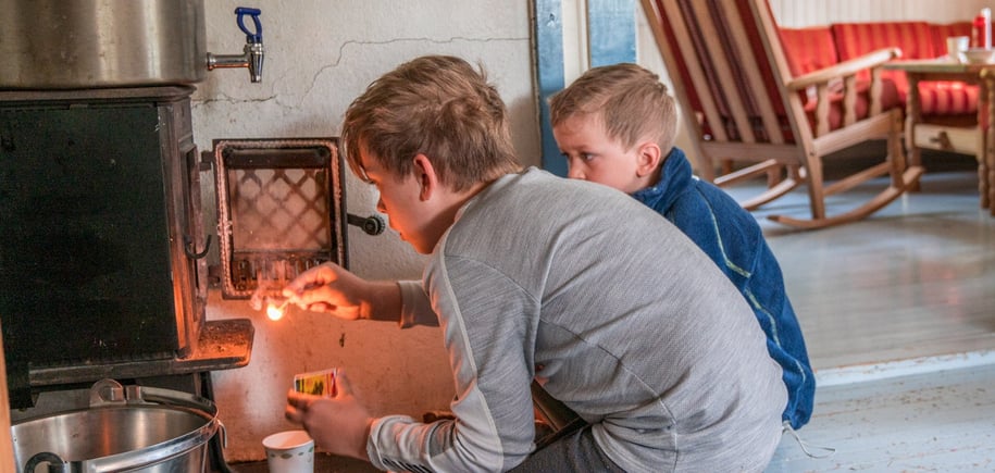 To gutter som fyrer i en vedovn. Den ene gutten har en fyrstikk i hånden.