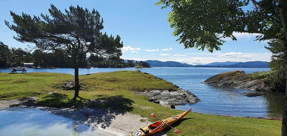 Kajakk ligger på sandstrand i idyllisk frilkuftsområde med sol, sjø og grønn gressplen.