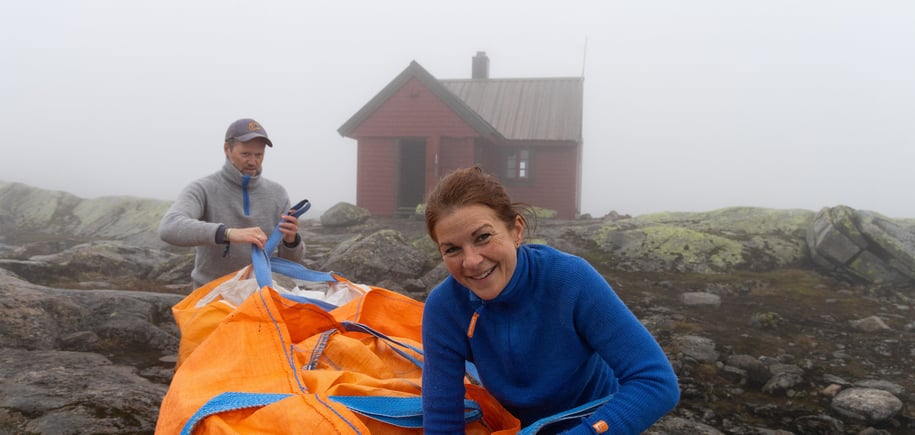 Kvinne i blå genser smiler foran hytte i tåkeheim.