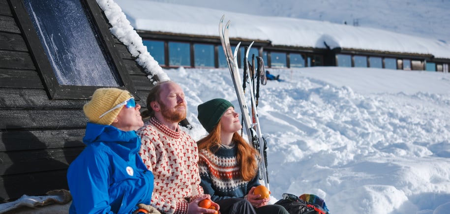 Tre personer sitter foran en lavo og nyter sola i snøen.