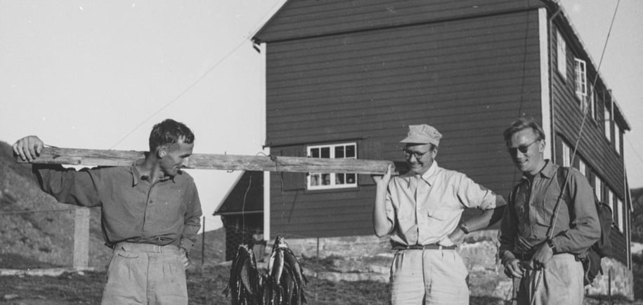 Gammelt bilde i svart-hvitt av tre menn som holder opp fisk foran hytte.