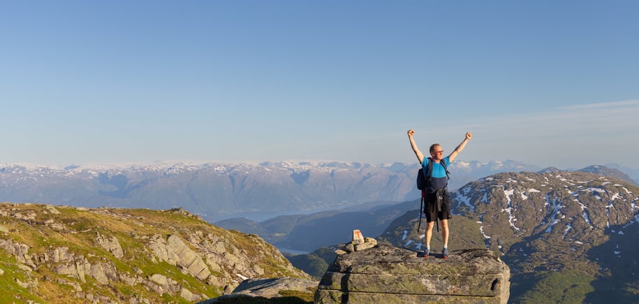 Mann 40+ står på stein med hendene i været, smiler, med Hardangerfjorden og fjell bak seg.