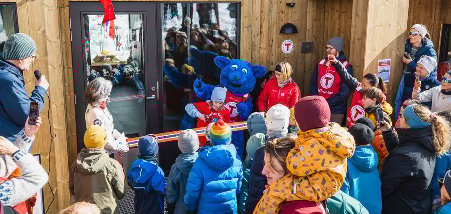 Ei jente klipper snoren foran døren på en ny og moderne hytte. Rundt henne står mange barn og voksne, og Barnas turlag sin maskot, den blå fjellreven Turbo.