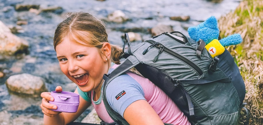 En liten jente sitter på huk og drikker fra en lilla kopp ved en elv. Hun smiler til kamera.