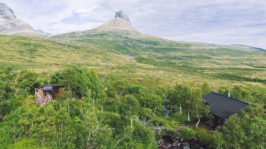 I forgrunnen to hytter som ligger i skogen med fjelltopp i bakgrunnen