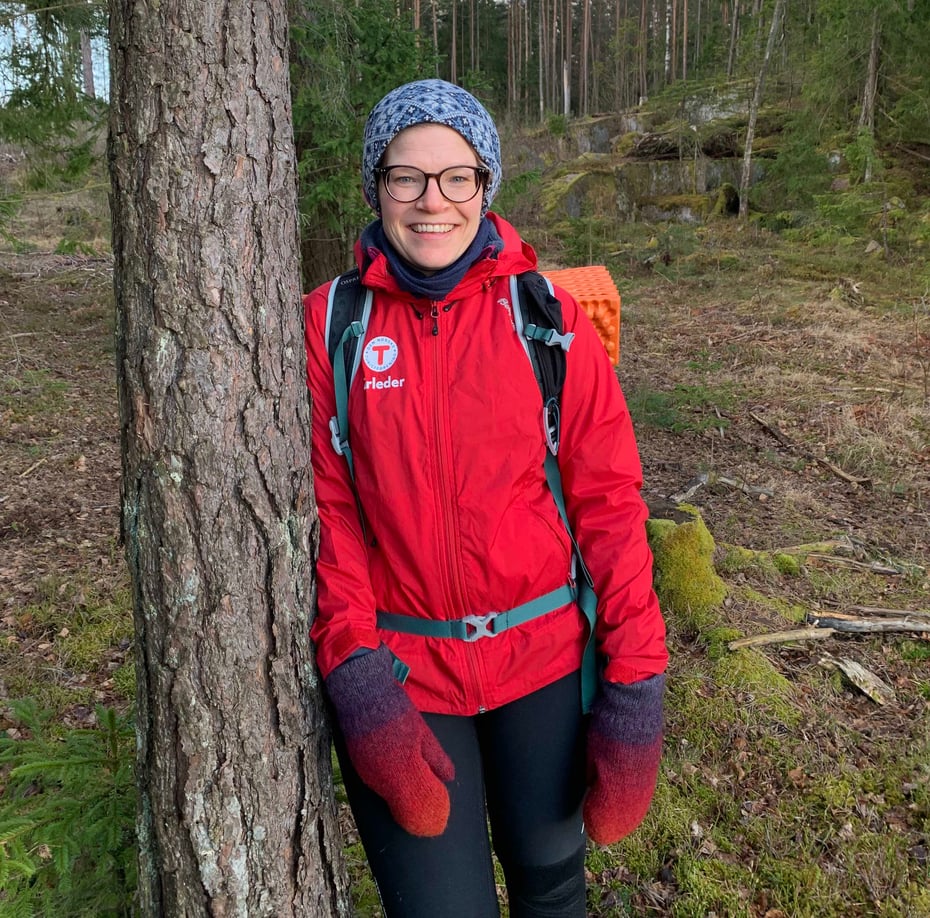 Smilende kvinne i rød DNT-jakke som lener seg mot et tre.