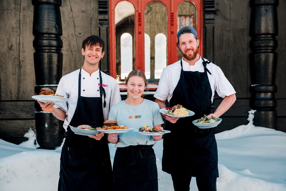 Foto. Tre perosner med forkle holder frem tallerker med mat. Gammlet trebygg i bakgrunnen