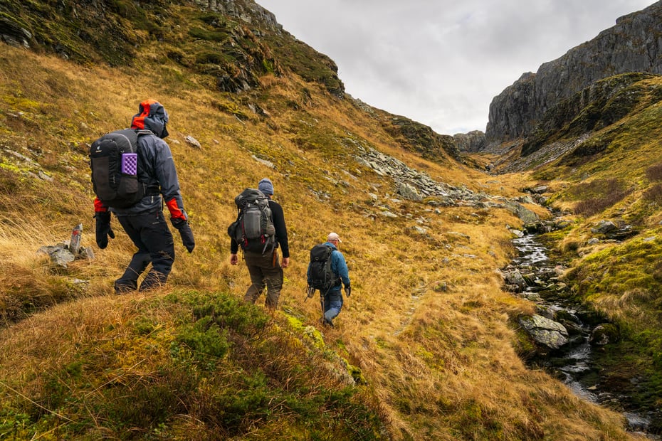 3 personer går tur med sekk. Det er høstfarger som brunt og grått i terrenget. De er godt kledd med votter og hetten oppe. Tydelig at det er vått